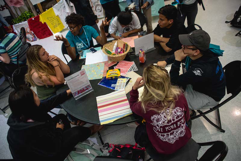 A group of 7 students sit and talk to each other around a table covered in markers, paper, and other crafting materials. 