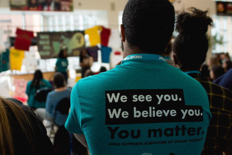 A person stands watching the Take Back the Night Speak Out. The back of their teal shirt says “We see you. We believe you. You matter.”