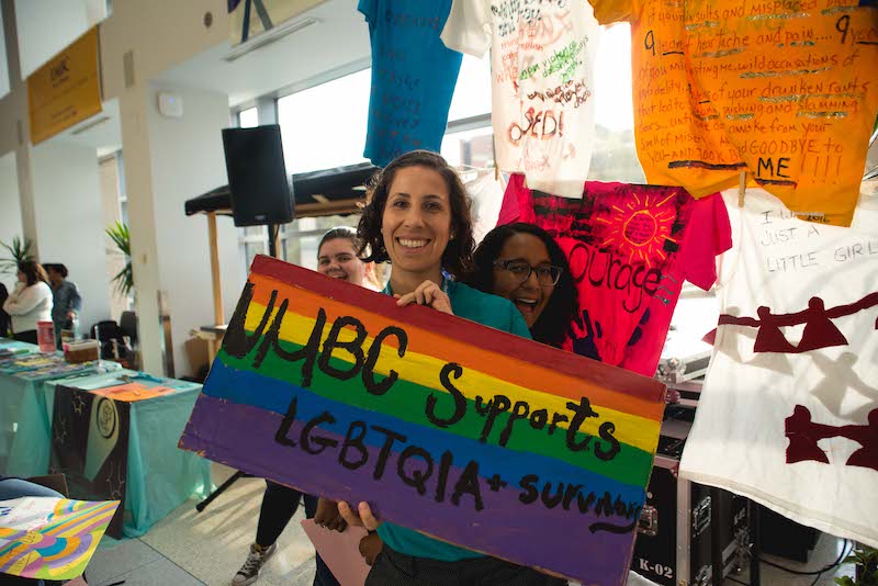Three people standing in front of Clothesline Project T-shirts. One of them is holding a sign that says “UMBC Supports LGBTQIA+ Survivors”