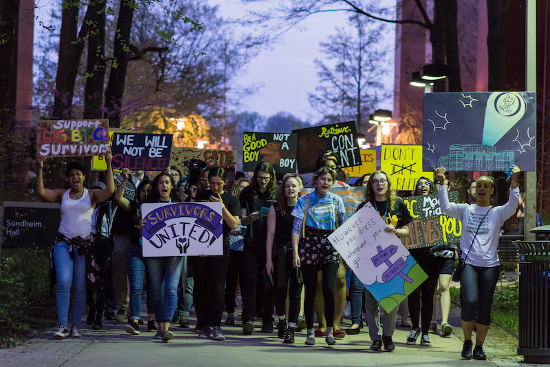 A head-on view of the Take Back the Night march as night falls with marchers on Academic Row holding signs with pro-consent messages.
