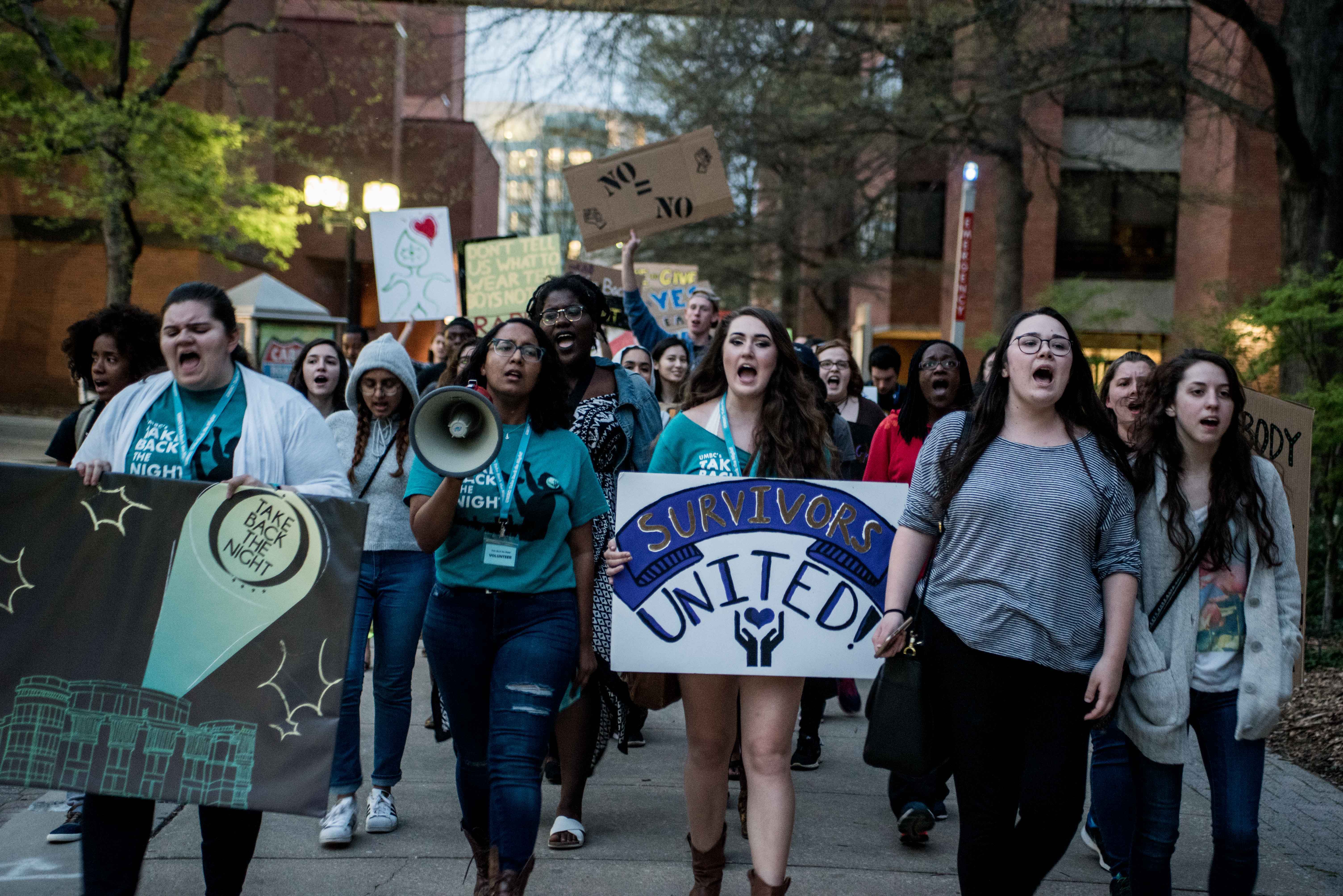 Five students are mid-chant at the front of the Take Back the Night march with a crowd behind them. One student holds a megaphone, one holds a sign that says “Survivors United!”